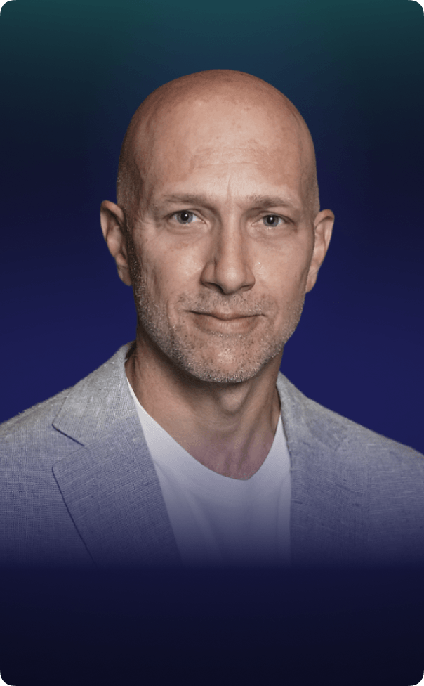 Gus Fritschie wearing a blue jacket over a white t-shirt stands confidently, smiling at the camera.